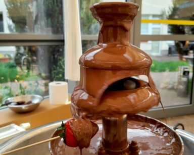 Zbliżenie. Garnek z fontanną z której wylewa się ciepła czekolada. Obok ,ktoś na patyczku macza w czekoladzie truskawkę.