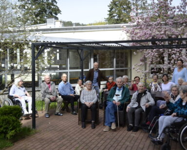 Ogród. Grupa seniorów i opiekunów siedzi na meblach ogrodowych. W tle drzewa magnolii pełne kwiatów różowych i białych.