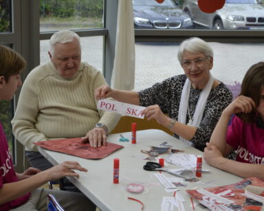 Zbliżenie. Dwoje seniorów i dwóch wolontariuszy siedzi przy stole. Seniorka podniosła napis POLSKA z orłem w koronie.