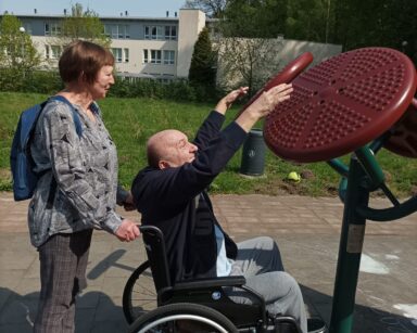 Zbliżenie. Kobieta stoi za wózkiem inwalidzkim, na wózku senior. Mężczyzna sięga rękoma do urządzenia do ćwiczeń.