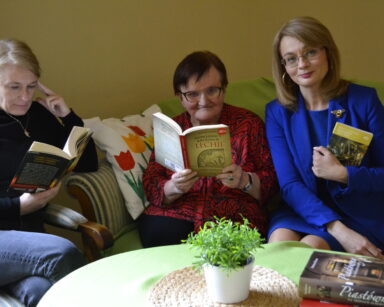 Zbliżenie. Na kanapie siedzą trzy kobiety, trzymają różne książki. Przed nimi stolik, na nim kwiat i książka