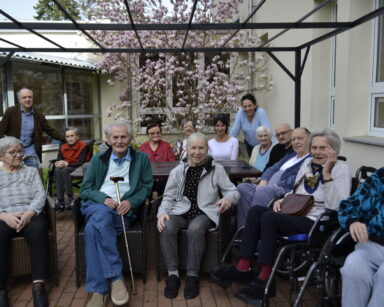 Ogród. Grupa seniorów i opiekunów siedzi przy stoliku. W tle rozłożyste drzewo magnolii o różowych kwiatach.