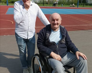 Zbliżenie. Boisko. Dwóch panów pozuje do zdjęcia. Jeden jest w jasnej koszuli trzyma wózek, senior na wózku ubrany w dres.