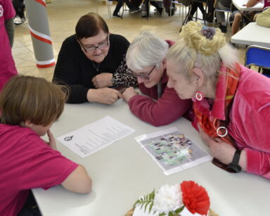 Zbliżenie. Trzy seniorki i młody wolontariusz pochylają się nad stołem, na którym leży test wiedzy z Polski.