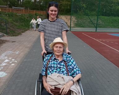 Zbliżenie. Boisko. Młoda kobieta prowadzi na wózku seniorkę. W tle dwie seniorki na spacerze.