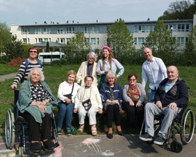 Zbliżenie. Grupa seniorów i opiekunów pozuje do zdjęcia. W tle trawa, budynek DPS, biały samochód.