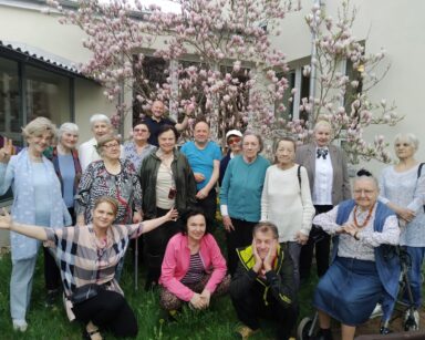 Ogród. Grupa uśmiechniętych seniorów pozuje do zdjęcia pod drzewem magnolii o różowych kwiatach.