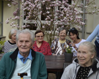 Ogród. Grupa seniorów i opiekunka siedzą przy stoliku. W tle drzewo magnolii.