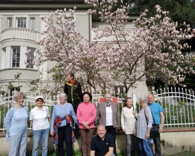 Grupa osób z DDP pozuje do zdjęcia. W tle biały ozdobny płot, drzewo magnolii, jasny zabytkowy dom.