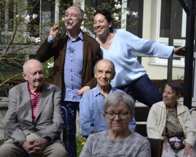Ogród. Grupa osób pozuje do zdjęcia. Mężczyzna wącha kwiat magnolii, obok niego uśmiecha się kobieta. Przy stoliku seniorzy.