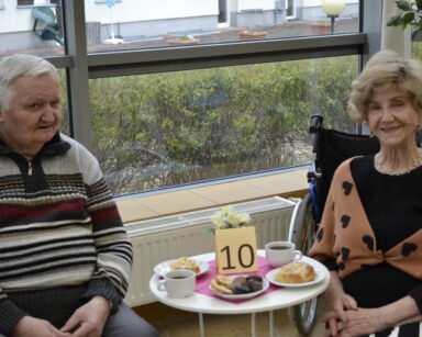 Zbliżenie. Para uśmiechniętych seniorów siedzi przy stoliku. Na stoliku nr 10, ciasteczka, herbata w filiżankach.