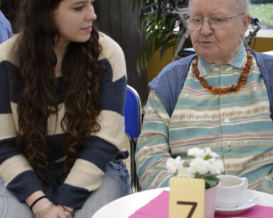 Zbliżenie. Seniorka siedzi z młodą dziewczyną przy stoliku. Na stoliku numer 7, ciastka, kwiat w donicy i filiżanka.
