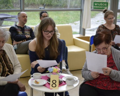 Zbliżenie. Trzy kobiety siedzą przy stoliku i trzymają tekst na kartkach. Kobiety śpiewają. W tle trzy osoby obserwują salę.