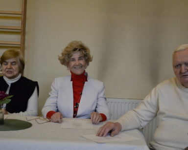 Zbliżenie. Troje uśmiechniętych seniorów siedzi przy stole. Na stole kartki z napisami. Wazon z purpurowym kwiatem.