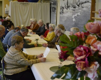 Sala. Grupa seniorów przy długim stole. Na stole wazony z kwiatami, przed seniorami kartki z tekstem. W tle wolontariusze.