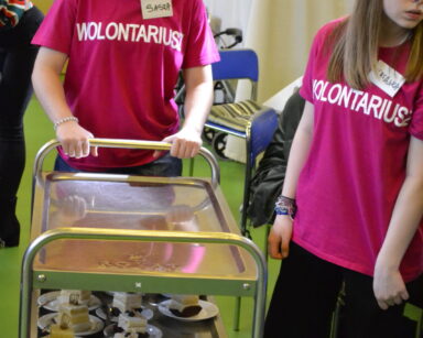 Zbliżenie. Dwie wolontariuszki z plakietkami Natalka i Sasza stoją przy wózku. Na nim na talerzykach ciasta.