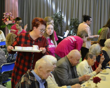Sala. Grupa ludzi. Przy stołach seniorzy, między stołami chodzą opiekunowie, wolontariusze. W tle rośliny, bukiety kwiatów.