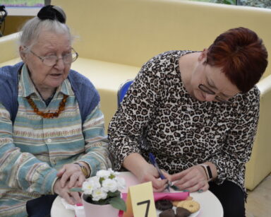 Zbliżenie. Kobieta w krótkich rudych włosach pochyla się nad stołem i pisze na kartce. Obok siedzi seniorka.