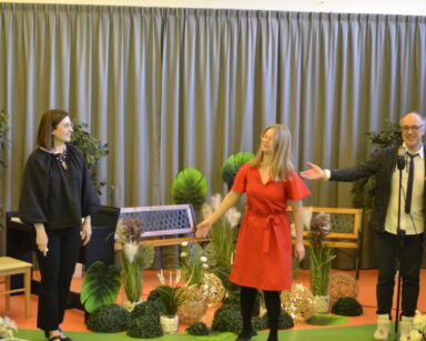 Scena. Dwie kobiety i mężczyzna przy mikrofonie. Na scenie dekoracje z zielonych roślin i lampek. Pomiędzy stoją ławki.
