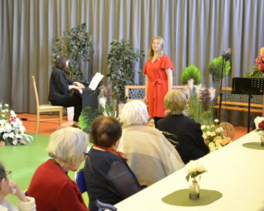 Sala. Dekoracje z zielonych roślin, kwiatów i światełek. Na scenie dwie kobiety, jedna przy pianinie. Przy stole seniorzy.