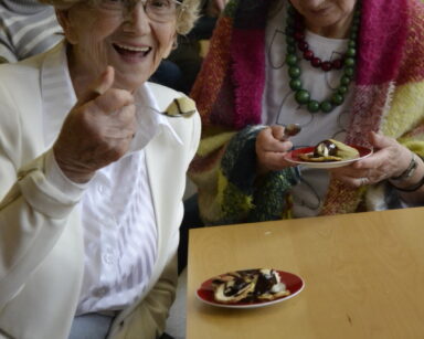 Zbliżenie. Dwie seniorki pozują do zdjęcia. Jedna kobieta w ręku trzyma łyżeczkę z owocami i czekoladą. Na stole deser.