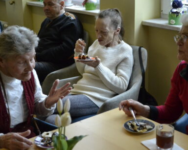 Zbliżenie. Trzy seniorki siedzą przy talerzykach pełnych owoców oblanych czekoladą. Na stoliku obok filiżanka z kawą.