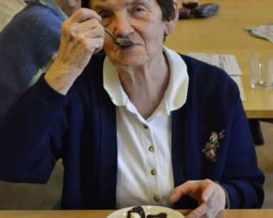 Zbliżenie. Seniorka pozuje do zdjęcia. Przy ustach trzyma łyżeczkę, na talerzu owocowy deser polany czekoladą.