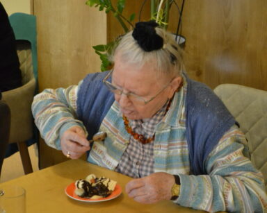 Zbliżenie. Seniorka siedzi przy stole. Bierze łyżkę do ust. Na talerzu przed nią owoce oblane czekoladą. W tle kwiat.
