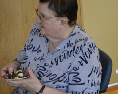 Zbliżenie. Seniorka w fioletowej bluzce w czarne szlaczki siedzi przy stole. W ręku trzyma deser oblany czekoladą.