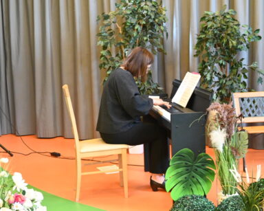 Zbliżenie. Przy pianinie kobieta w czerni. Na pianinie nuty. Obok ławka parkowa. Dookoła dekoracje z kolorowych roślin.