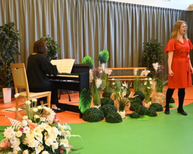 Zbliżenie. Scena. Dwie kobiety. Jedna śpiewa, druga gra na pianinie. Dookoła roślinne dekoracje i parkowa ławeczka.