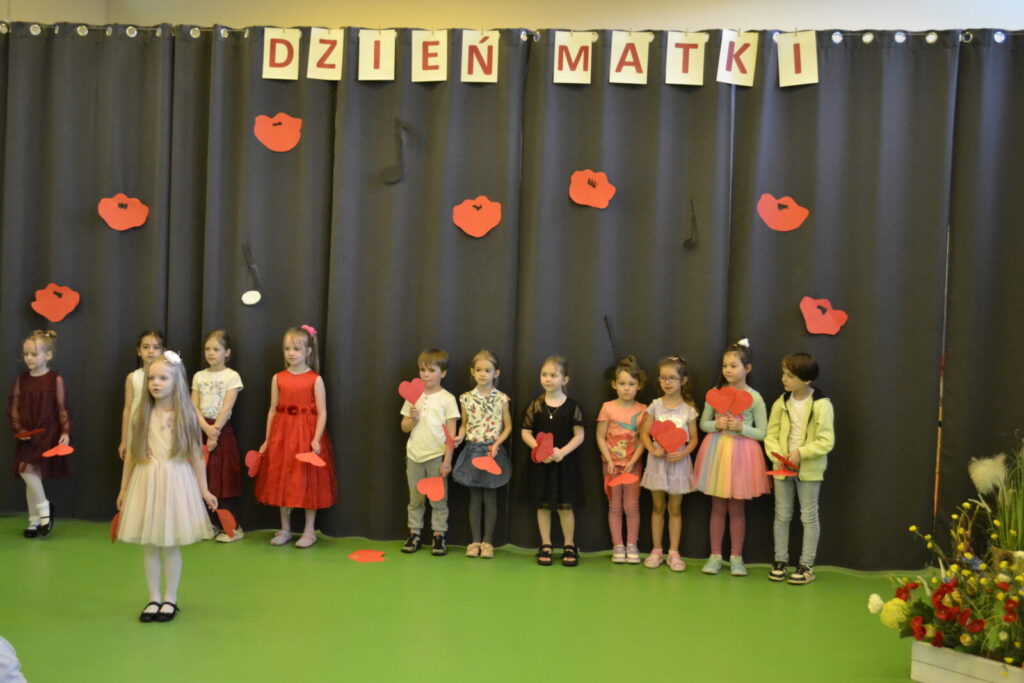 Sala. Scena. Grupa przedszkolaków w kolorowych ubrankach. W rączkach trzymają serduszka. W tle napis "Dzień Matki".