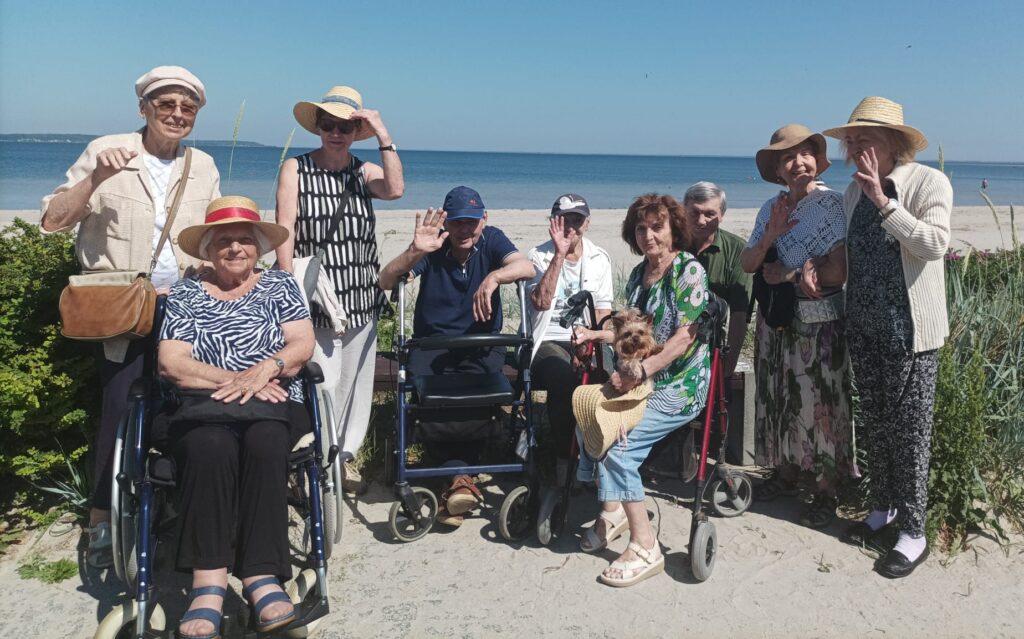 Plaża. Grupa seniorów pozuje do zdjęcia. Seniorzy mają na głowach kapelusze i czapkami z daszkiem. W tle piasek i morze.