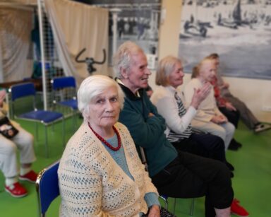 Zbliżenie. Seniorka w ażurowym swetrze patrzy w stronę fotografa, pozuje do zdjęcia. Obok siedzą seniorzy.