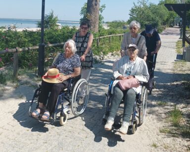 Plaża. Promenadą spacerują seniorzy, dwie seniorki pchają wózki, obok senior na chodziku. W tle róże, piach, morze, łodzie.