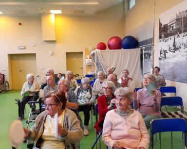 Sala. Grupa seniorów siedzi na krzesłach. w dłoniach trzymają papierowe talerze. W tle zdjęcia na ścianach.