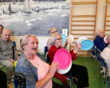 Sala. Uśmiechnięte seniorki patrzą na scenę. W dłoniach trzymają kolorowe talerze papierowe.