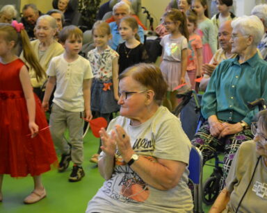 Sala. Grupa dzieci przechodzi między siedzącymi na krzesłach seniorami. Seniorzy klaszczą, dzieci uśmiechają się.