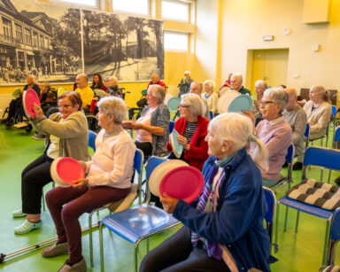 Sala. Grupa seniorów podnosi do góry dłonie. W dłoniach trzymają kolorowe talerze z papieru.