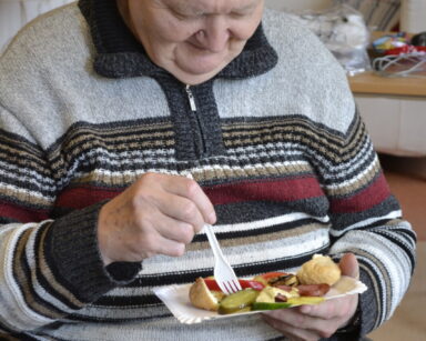 Zbliżenie. Senior w swetrze w paski siedzi na krześle i trzyma w ręku tackę z grillowanymi warzywami i kawałkiem chleba.