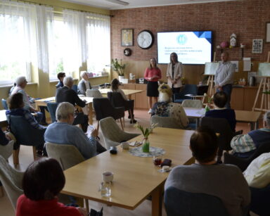 Sala. Grupa seniorów siedzi przy stole, patrzą w stronę 3 stojących ludzi przy monitorze. Na nim napis 