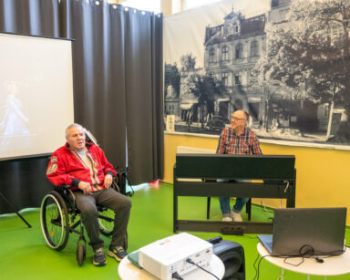 Sala. Scena. Senior na wózku śpiewa. Obok mężczyzna gra na pianinie.