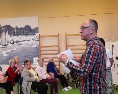 Sala. Mężczyzna z kartką w ręku stoi przodem do siedzących na krzesłach seniorów. Wszyscy patrzą na mężczyznę.