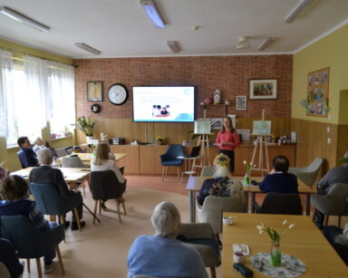 Sala. Grupa osób siedzi przy stołach. W tle kobieta przy plakatach z napisem 