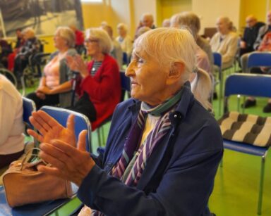 Sala. Seniorka w granatowej marynarce klaszcze w dłonie. W tle seniorzy również klaszczą w dłonie. Wszyscy uśmiechają się.