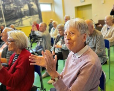 Sala. Seniorka w różowym sweterku klaszcze w dłonie i patrzy w stronę fotografa. W tle seniorzy również klaszczą.