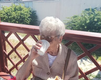 Zbliżenie. Seniorka z kubeczkiem przy ustach i bułką w ręce siedzi w altance. W tle budynek i zielone krzewy.