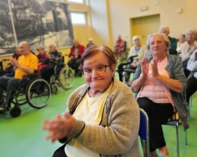 Sala. Uśmiechnięta seniorka w kremowym swetrze i opasce na głowie klaszcze w dłonie i pozuje do zdjęcia. W tle seniorzy.