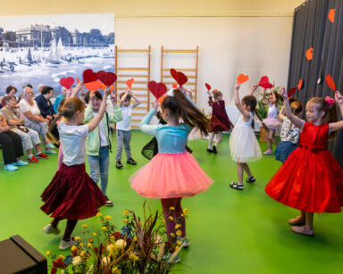 Sala. Grupa seniorów ogląda występ maluchów. Dzieci na scenie kręcą się w kółku unosząc do góry serduszka.