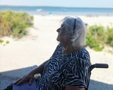 Zbliżenie. Seniorka siedzi na wózku, w rękach trzyma tablet. Patrzy gdzieś w dal. W tle trawy na piasku i morze.